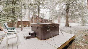 Een houten terras met een jacuzzi, een overdekte zithoek en een rustieke chalet in een besneeuwd bos.