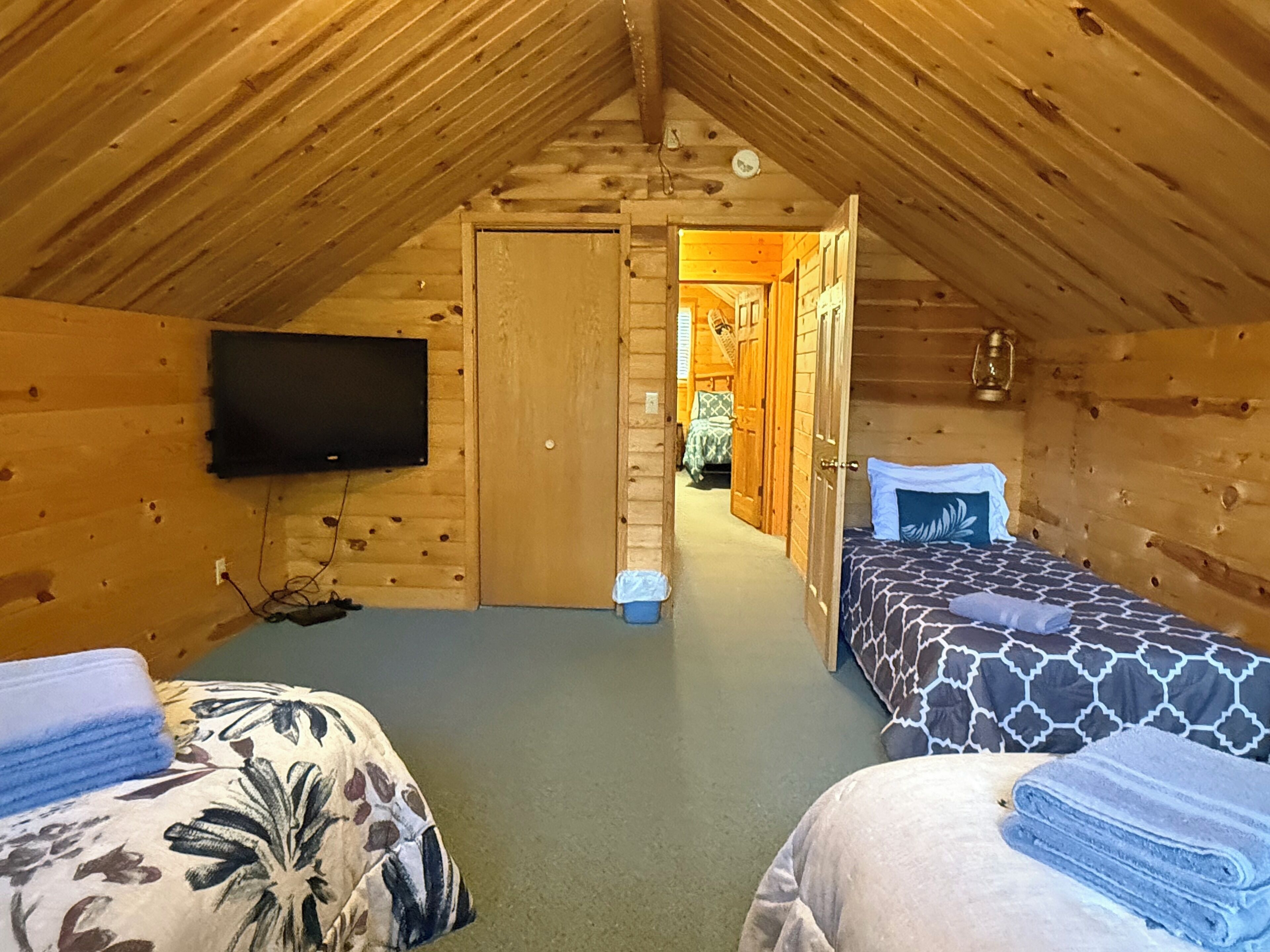 A cozy attic bedroom with two beds, a television, and a wooden ceiling.