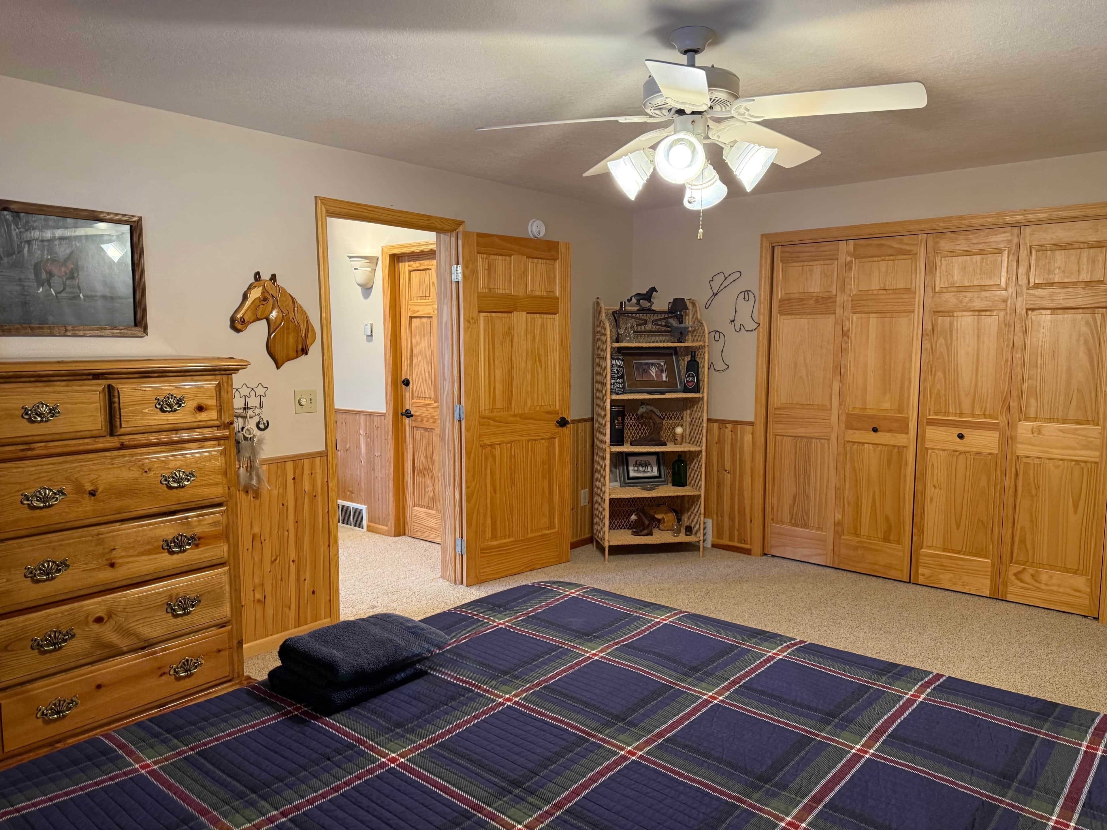 Une chambre à coucher comprenant un lit, une armoire en bois, une commode, un ventilateur de plafond et une étagère fixée au mur, sur laquelle sont disposés divers objets.