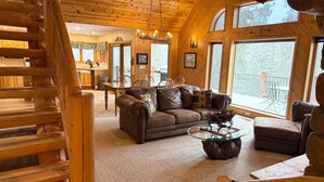 Ein gemütliches Blockhaus-Interieur mit einer Holztreppe, einem Wohnzimmer mit Ledersofa, einem Essbereich und großen Fenstern mit Blick ins Freie.