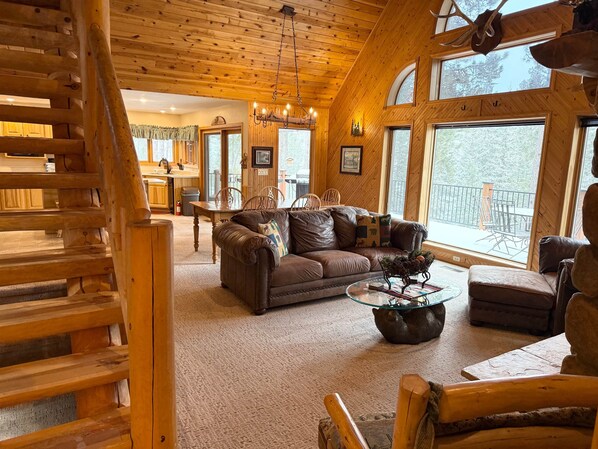 A cozy cabin interior with a wooden staircase, a living room with a leather sofa, a dining area, and large windows offering views of the outdoors.