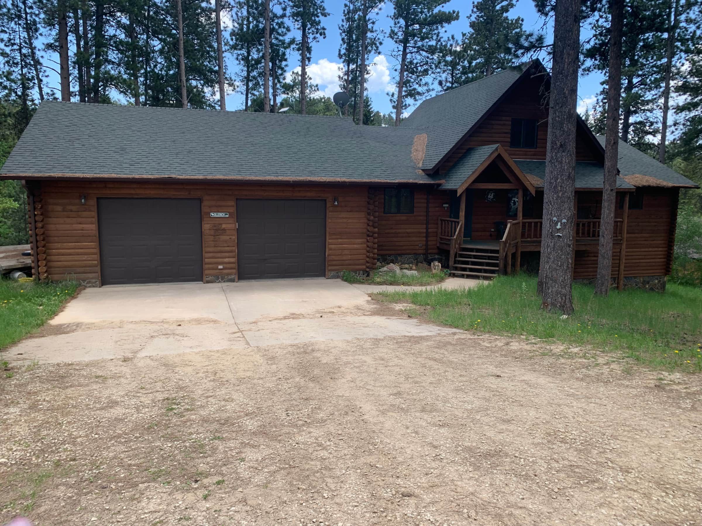 A log cabin with two garage doors, a front porch, and a dirt driveway.