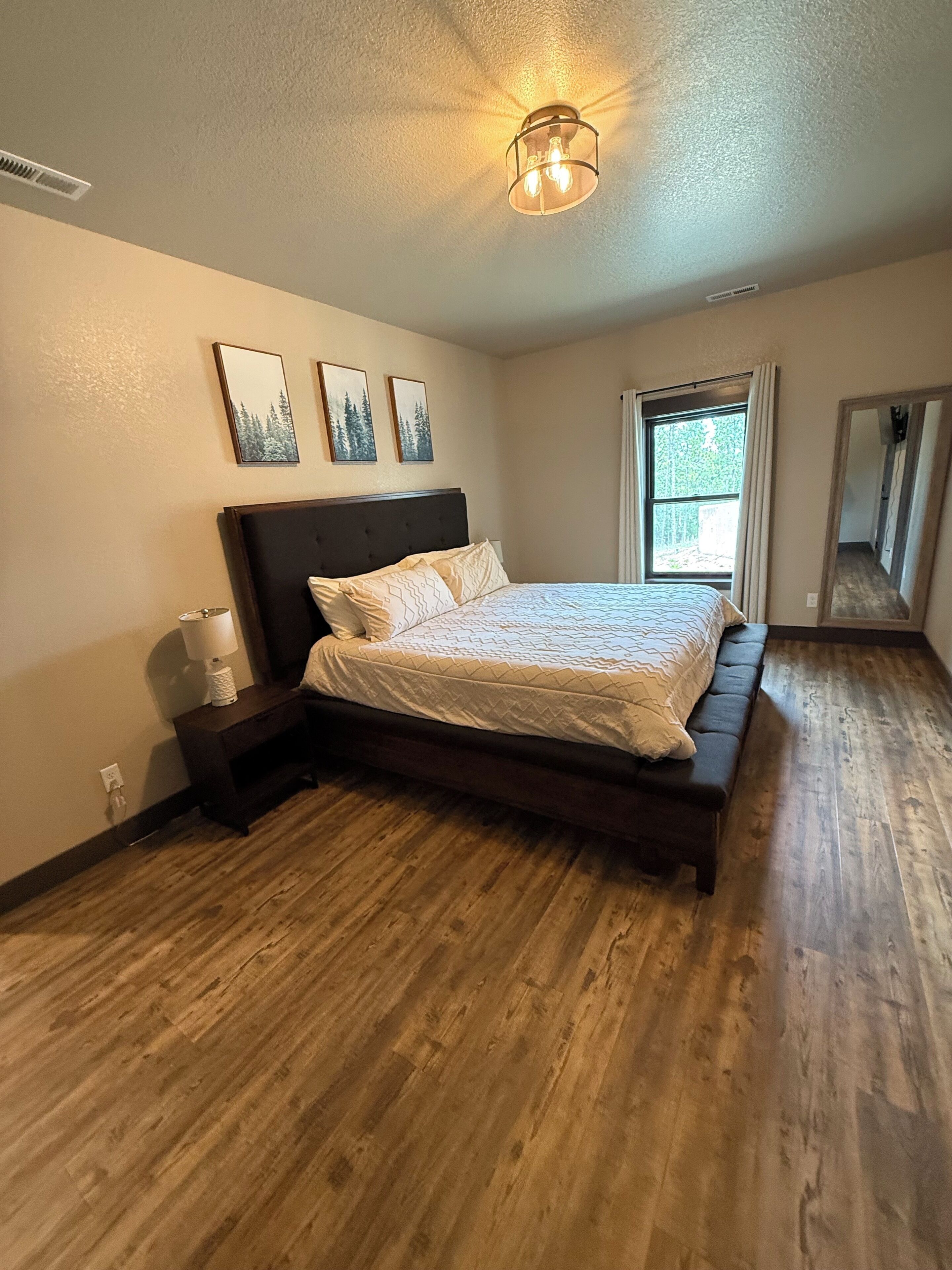 Una camera da letto con pavimento in legno, un letto con testiera, un comodino con una lampada e una finestra con tende.