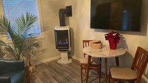 Een gezellige woonkamer met een houtkachel, een ronde eettafel met stoelen, een aan de muur bevestigde televisie en een potplant.