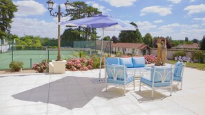 Un patio al aire libre con cancha de tenis, sillas de metal blanco, una mesa y un paraguas morado.