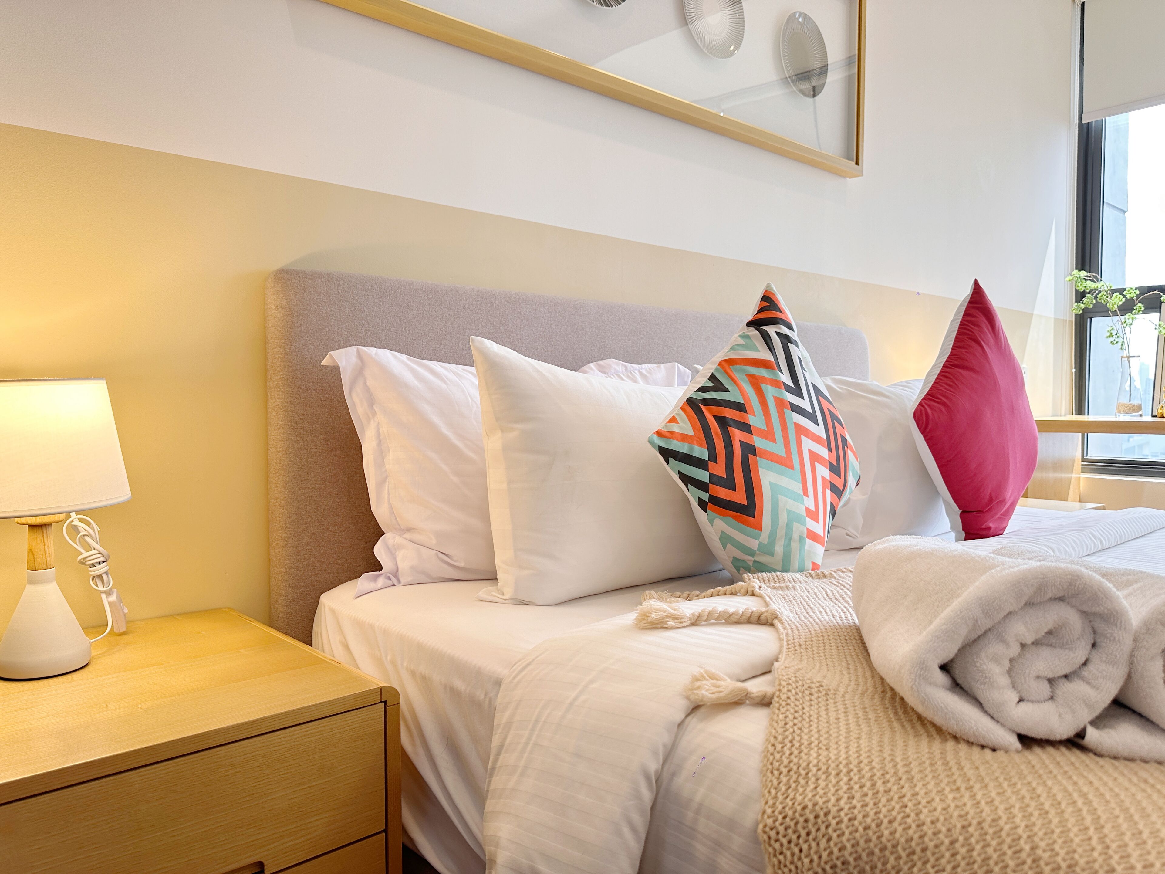 A neatly made bed with white bedding, a colorful geometric pillow, and a rolled-up towel.
