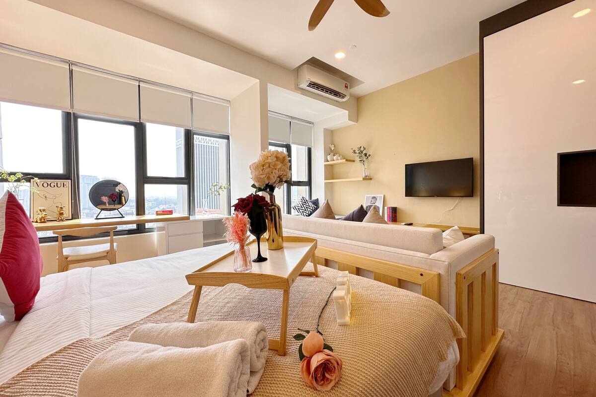 Quarto de hotel moderno com uma cama grande, uma escrivaninha de madeira, um sofá e uma televisão fixada na parede.