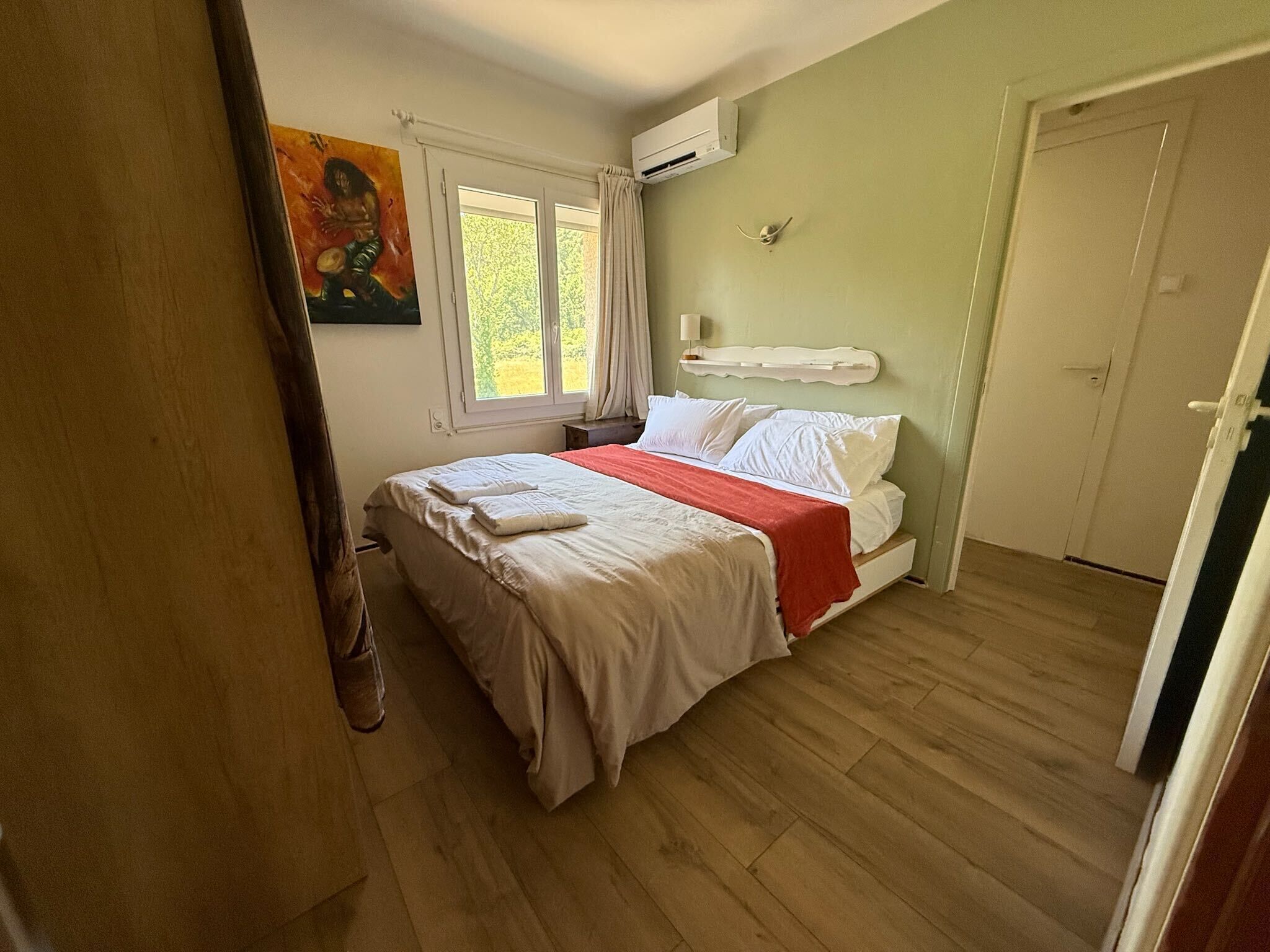 A bedroom with a bed, a window, an air conditioning unit, and a door.