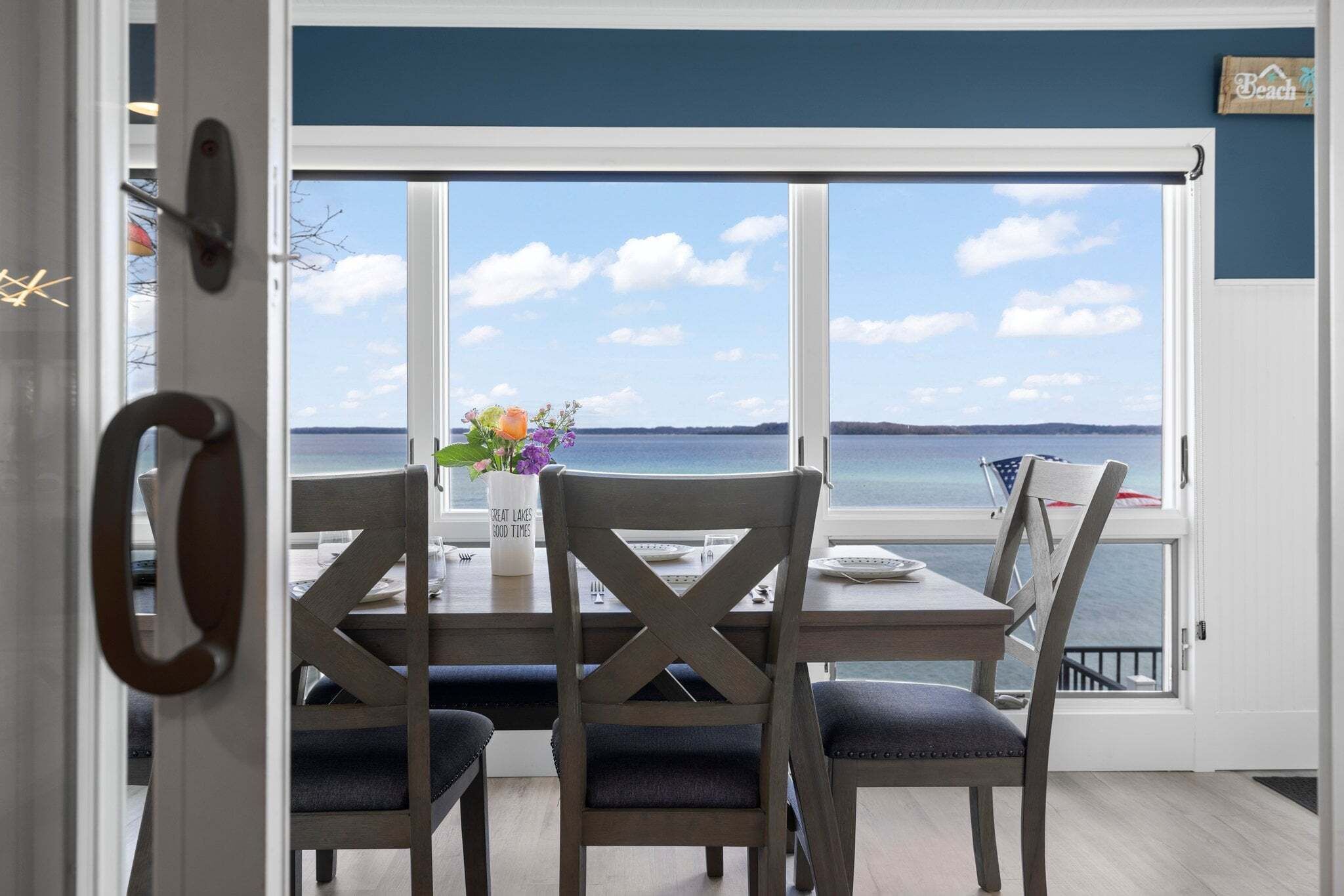 A dining area with a table and chairs, a vase with flowers, and a view of the water through large windows.