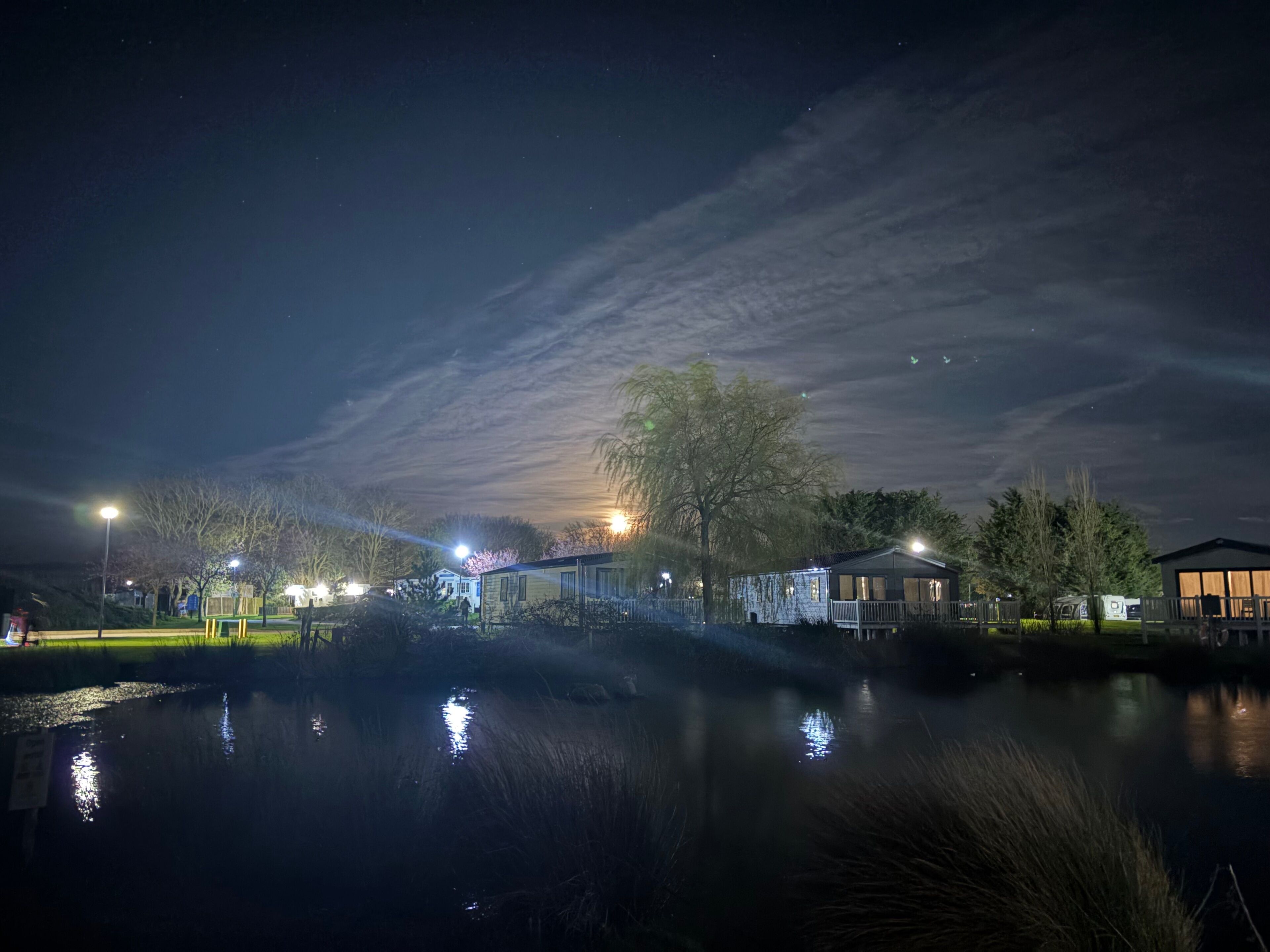 A nighttime view of a lakeside area with buildings, a dock, and a reflection on the water.