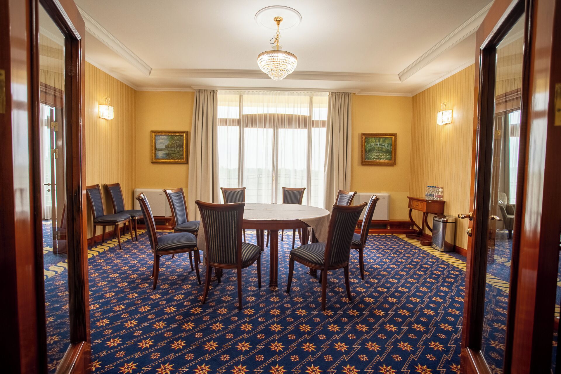 A conference room with a round table, six chairs, a chandelier, and two paintings on the walls.