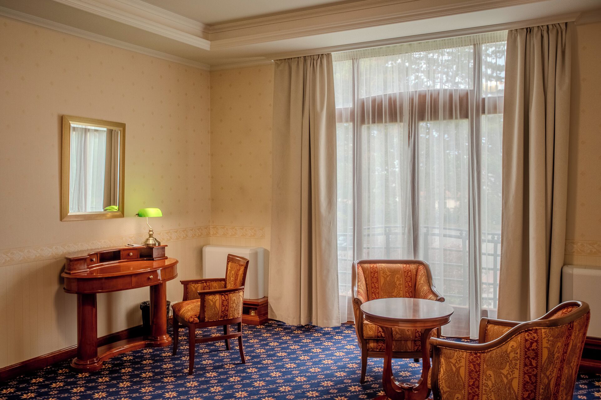 Ein Hotelzimmer mit Schreibtisch aus Holz, einem Sessel, einem kleinen Tisch, einem Spiegel und einem Fenster mit transparenten Vorhängen.
