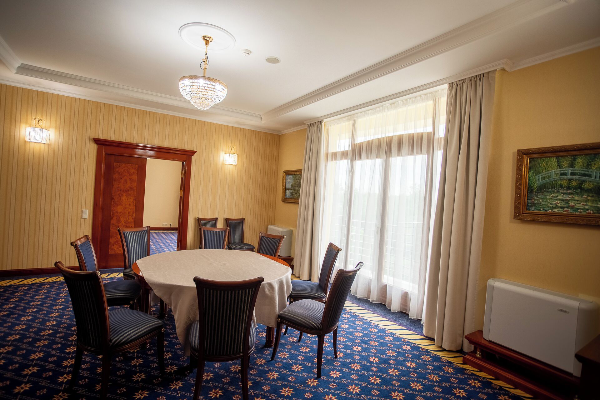 A conference room with a round table, six chairs, a chandelier, a painting, and a radiator.