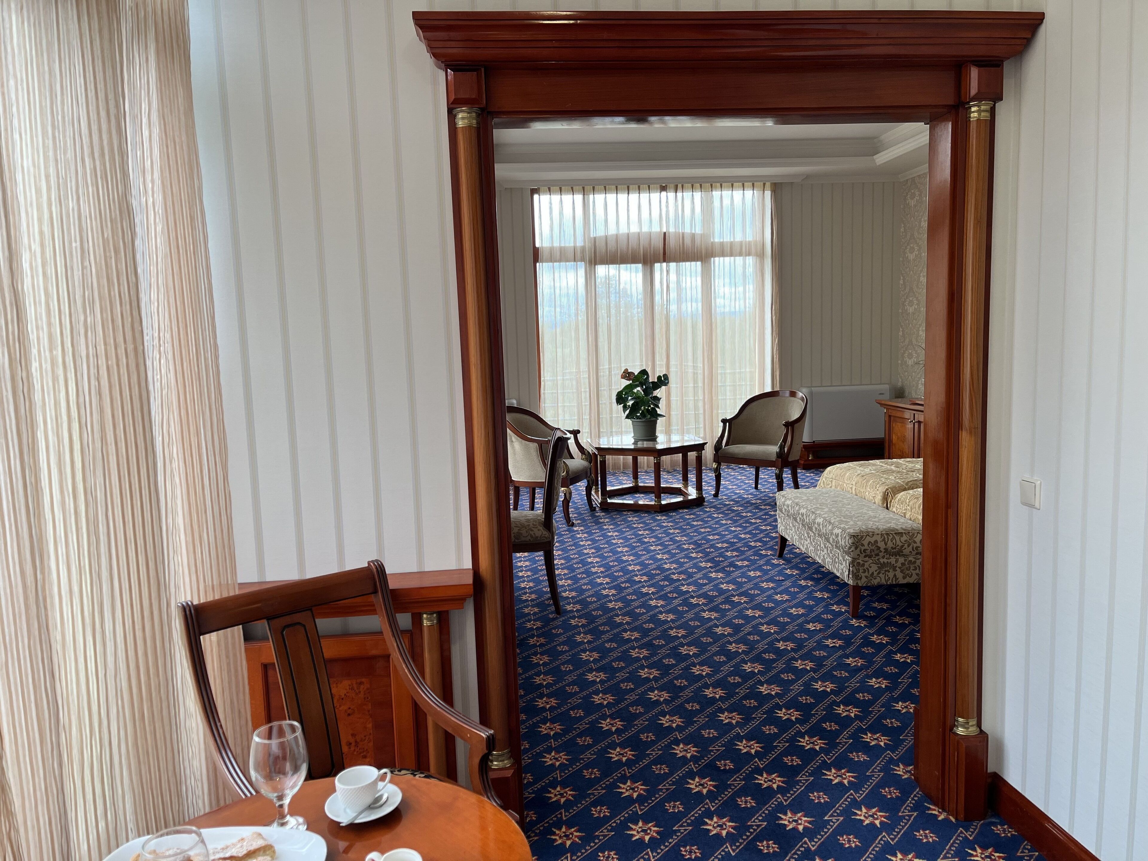 Habitación de hotel con alfombra azul estampada, una mesa de comedor de madera con sillas, una mesita redonda con una planta y una cama con colcha estampada.