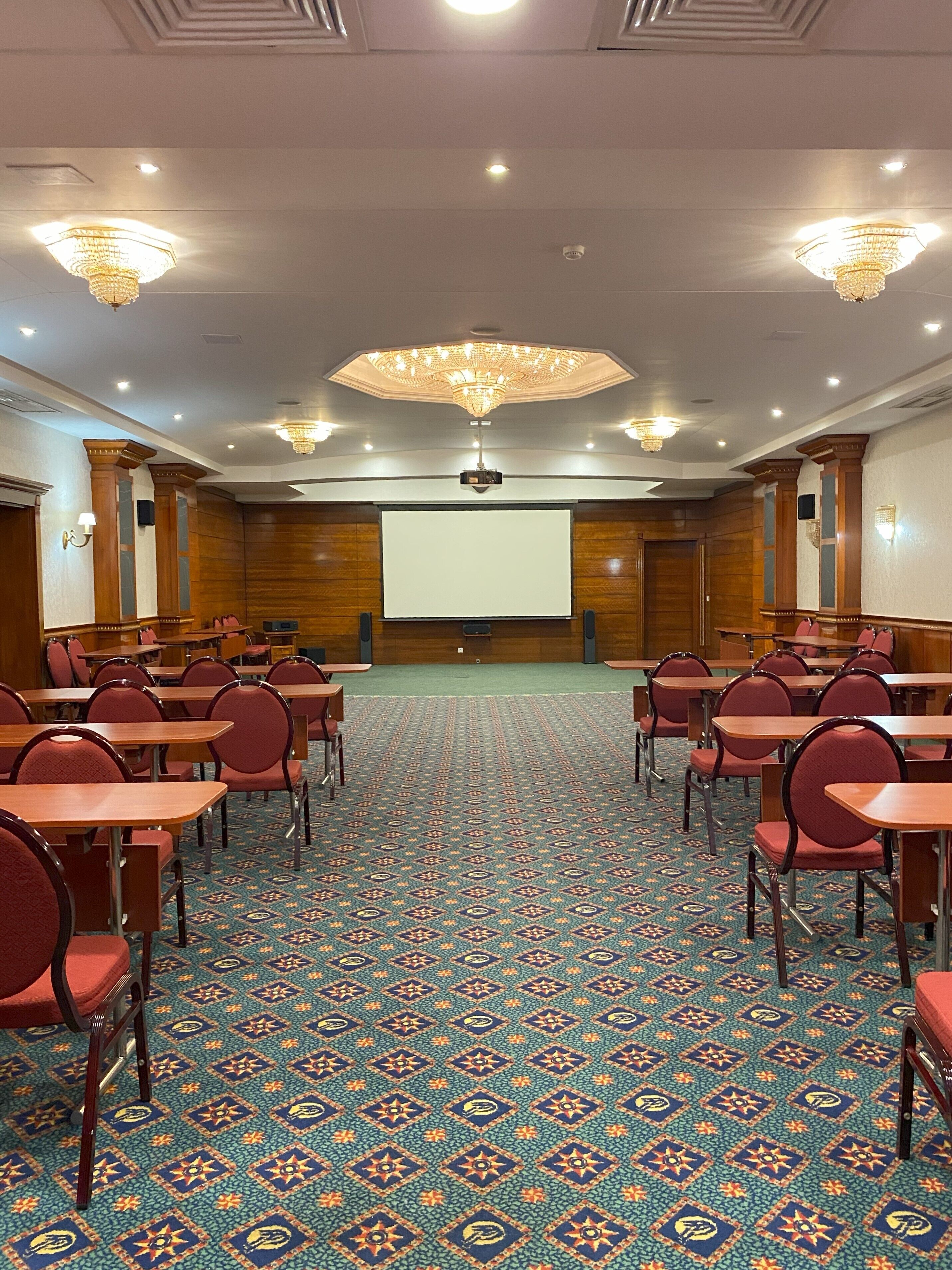 A conference room with a large screen, rows of tables and chairs, patterned carpet, and chandeliers.