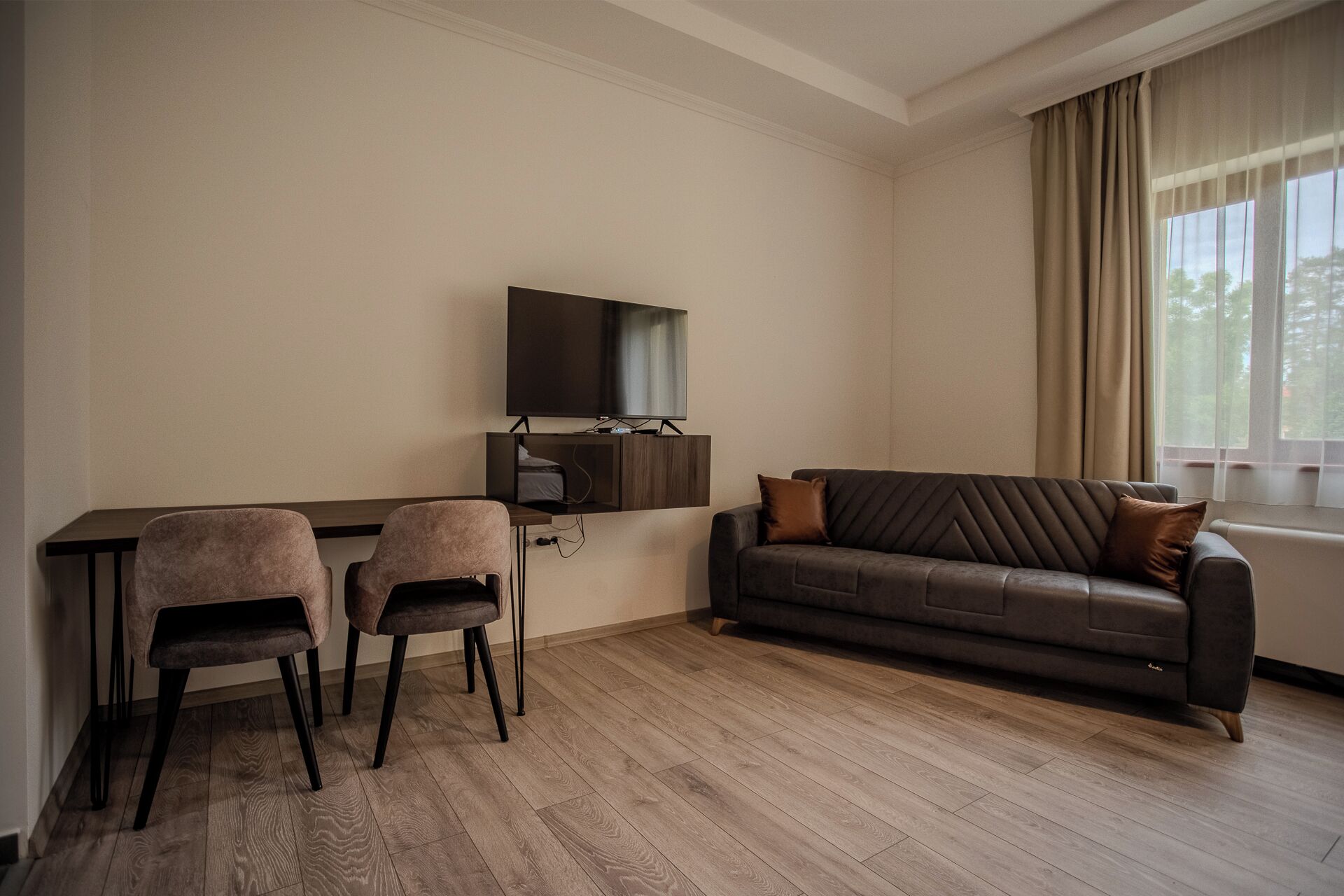 A living room with a dark brown leather sofa, two matching armchairs, a wooden coffee table, a flat-screen TV mounted on a wooden cabinet, and a large window with beige curtains.