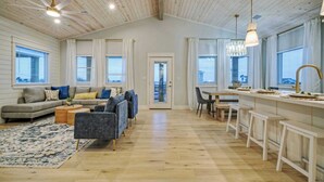 Un vaste espace de vie doté d’un plafond en bois, d’un ensemble de canapés, d’une table à manger avec des tabourets de bar et de grandes fenêtres avec des rideaux.
