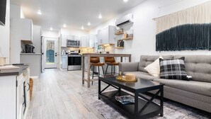 Un salon moderne avec un canapé gris, une table basse et un coin repas, la cuisine étant visible au fond.