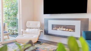 A modern living room with a fireplace, a white leather chair, and a TV mounted on the wall. - Zanjero by Avantstay Mins From Downtown Palm Springs Private Terrace, Shared Pool (Palm Springs)