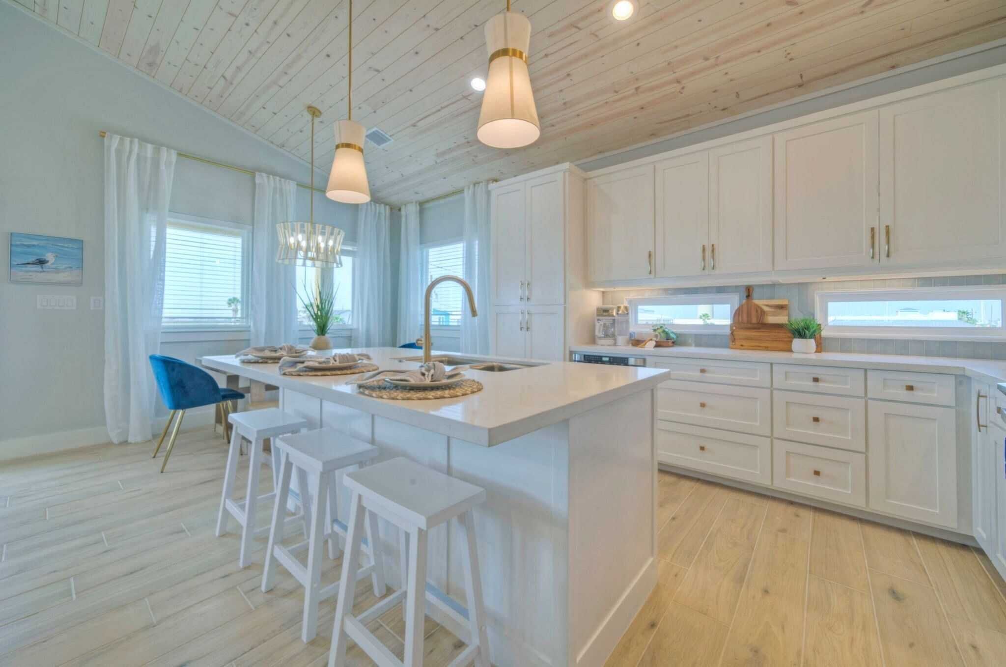Une cuisine moderne avec des meubles blancs, un îlot central doté de tabourets, un éclairage suspendu et une vue sur l’extérieur grâce à de grandes fenêtres.