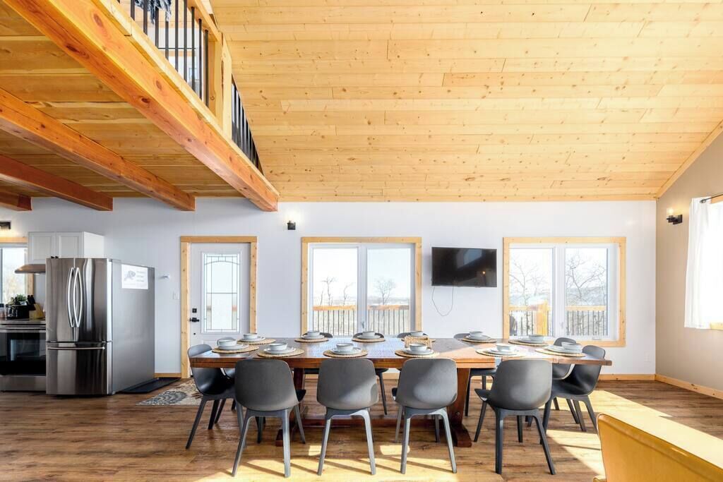 Un vaste espace dédié à la salle à manger, avec une longue table en bois pouvant accueillir six personnes, entourée de chaises modernes. Une grande fenêtre laisse entrer la lumière naturelle, et un téléviseur à écran plat est fixé au mur.