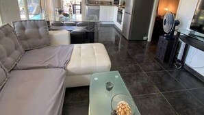 A modern living room with a grey sofa, a glass coffee table with decorative items, and a kitchen in the background.