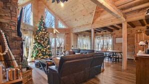 Ein gemütliches Blockhaus-Interieur mit einer großen Holzbalkendecke, einem Steinkamin, einem Weihnachtsbaum, einem Ledersofa und einem Essbereich.