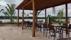 Een terras met tafels en stoelen, een houten pergola en uitzicht op een meer en groen.
