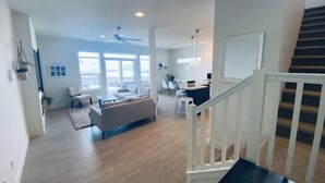 Ein geräumiger Wohnbereich mit einer Couch, einem Esstisch und Stühlen, einer Treppe mit Geländer und großen Fenstern für viel Tageslicht.