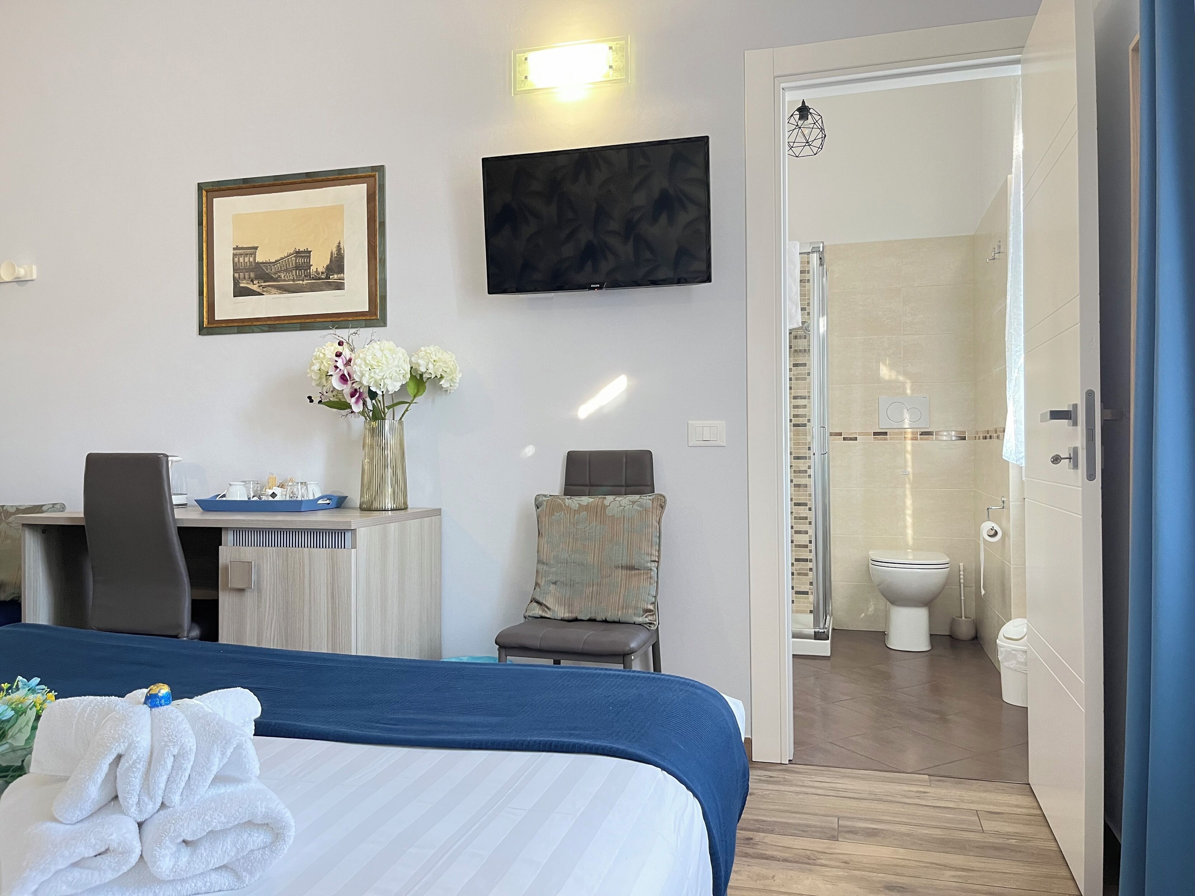 A hotel room with a bed, a desk, a chair, a TV, a framed picture, and a bathroom visible through an open door.