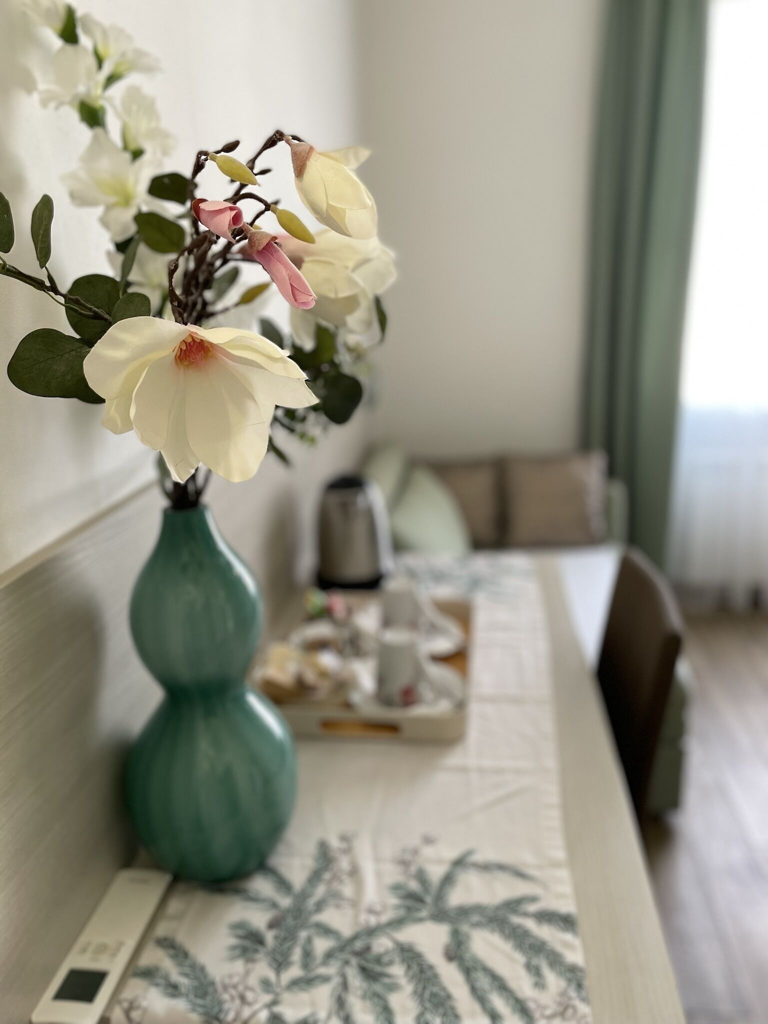 Una habitación con una cama, una mesita de noche con objetos sobre una bandeja y un jarrón con flores.