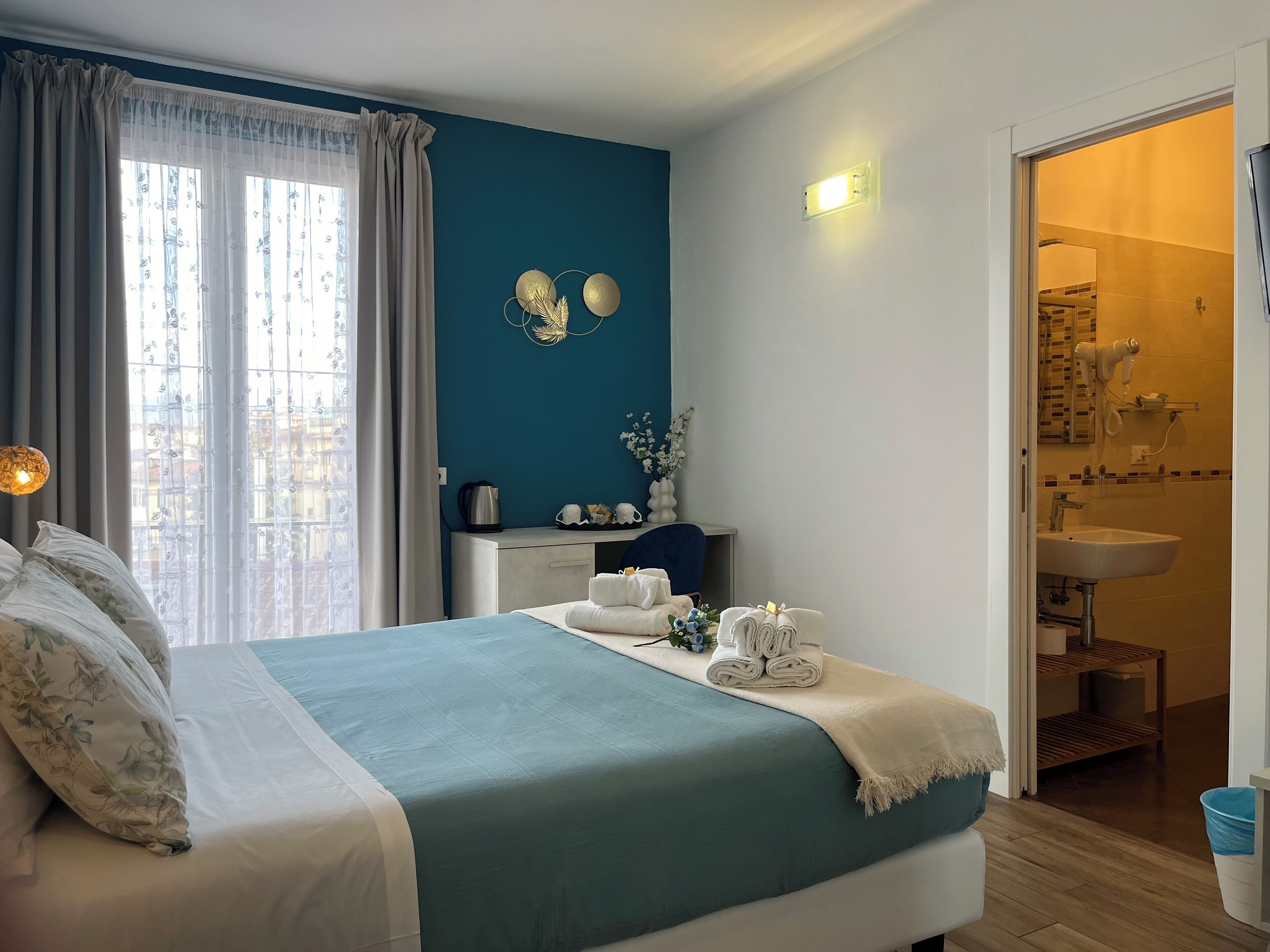 Une chambre à coucher avec un lit, une table de chevet sur laquelle se trouve une bouilloire, et une salle de bain visible à travers une porte ouverte.