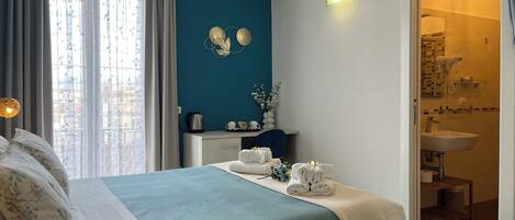 Une chambre à coucher avec un lit, une table de chevet sur laquelle se trouve une bouilloire, et une salle de bain visible à travers une porte ouverte.