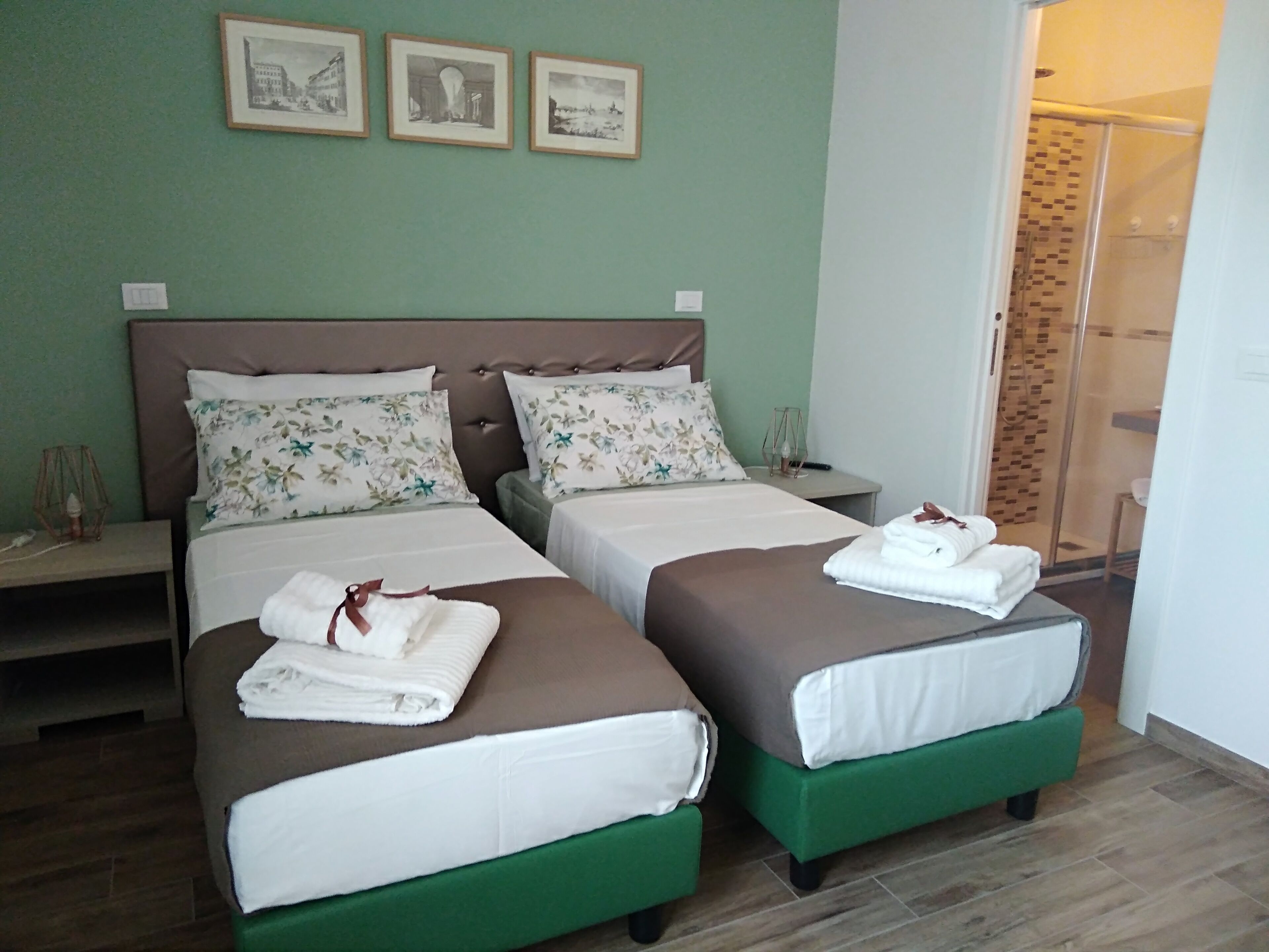 A hotel room with two beds, each with a green footboard, white bedding, and floral pillows. There are framed pictures on the wall and a glass shower door.