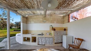 A covered outdoor kitchen area with a sink, stove, and refrigerator, a wooden chair, and a large woven bowl on a table. - 3745 Trullo Puspo by Barbarhouse (Carovigno)