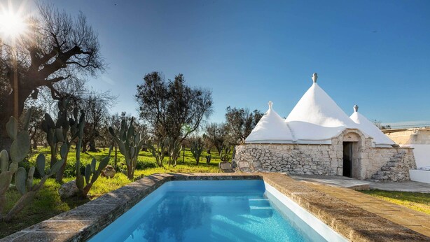A swimming pool with a stone edge, surrounded by cacti and trees, and a white building with conical roofs. - 3745 Trullo Puspo by Barbarhouse (Carovigno)