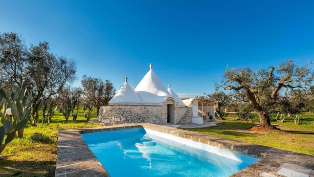 A swimming pool with a stone deck, a white building with a conical roof, and cacti in the background. - 3745 Trullo Puspo by Barbarhouse (Carovigno)