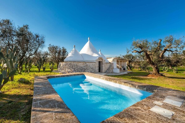 Una piscina con terraza de piedra, un edificio blanco con techo cónico y cactus al fondo.