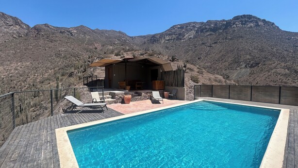 A rectangular swimming pool with a wooden deck, surrounded by a fence, and a patio area with lounge chairs and a table. - Elqui Valley Avocado Farm, 2-bedroom cabin in fabulous Vicuña with WiFi (Vicuña)