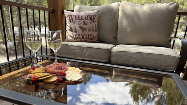 Een tafel met glazen blad, met een kaasplank, wijnglazen en een kussen met de tekst 'Welcome to the Woods' op een bank.