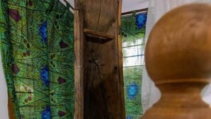 A wooden chair in a room with a peacock-patterned curtain and a window with a stained glass design. - Nazi - Explore semplicity, savor local cousine, immerse yourself in nature  (Michamvi Kae)