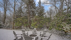 Een terras buiten bedekt met sneeuw, met Adirondack-stoelen en een vuurkorf, omringd door bomen en struiken.