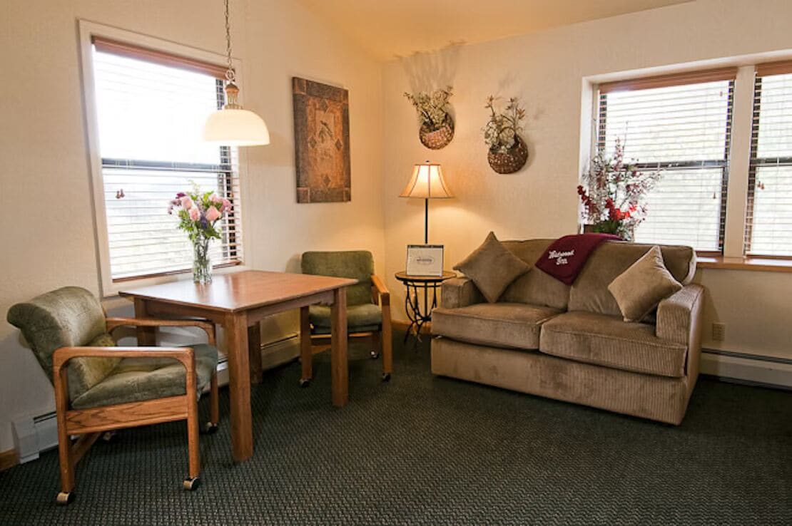 A cozy living room with a sofa, a wooden table, and two chairs. There is a lamp on a side table and a vase with flowers on the table.