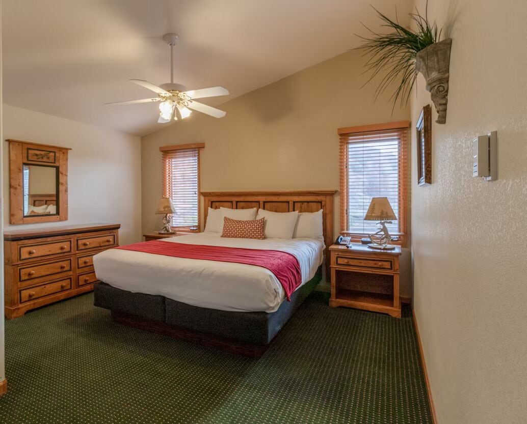 A bedroom with a large bed, wooden headboard, a ceiling fan, a nightstand with a lamp, and a mirror on the dresser.