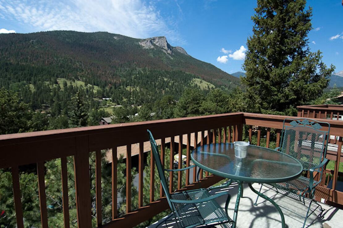 A balcony with a glass table and metal chairs, overlooking a mountainous landscape.