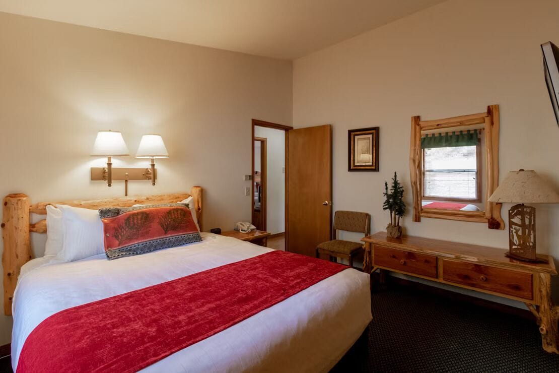 A hotel room with a large bed, a wooden headboard, two wall-mounted lamps, a small desk with a lamp, a chair, and a window with a curtain.