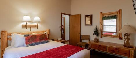 A hotel room with a large bed, a wooden headboard, two wall-mounted lamps, a small desk with a lamp, a chair, and a window with a curtain.