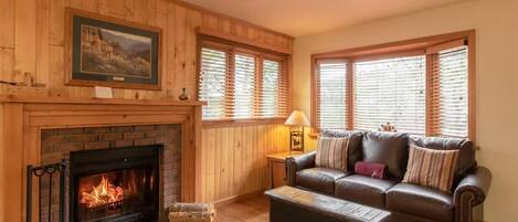 Un salon chaleureux avec une cheminée, des murs en bois, un canapé en cuir et une fenêtre avec des stores.