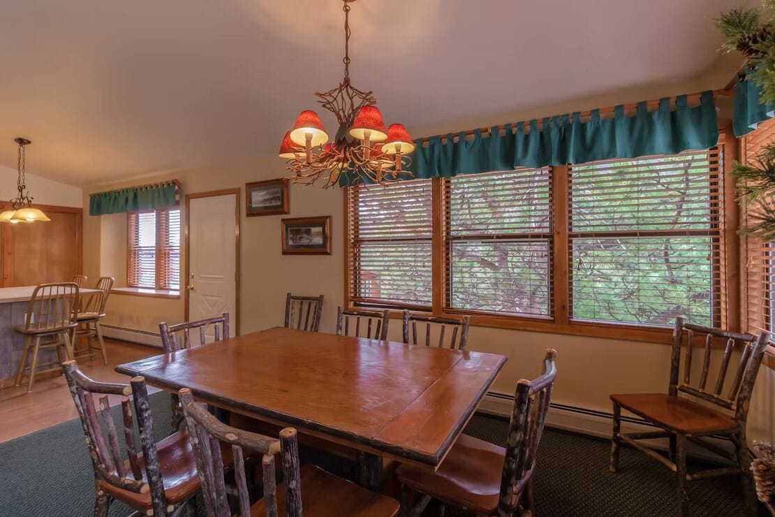A dining area with a wooden table and chairs, a chandelier, and windows with blinds.