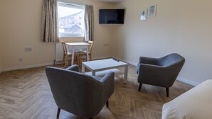 A modern living room with a herringbone wood floor, a grey armchair, a white coffee table, and a television mounted on the wall. - Wick Bay Apartments (Wick)